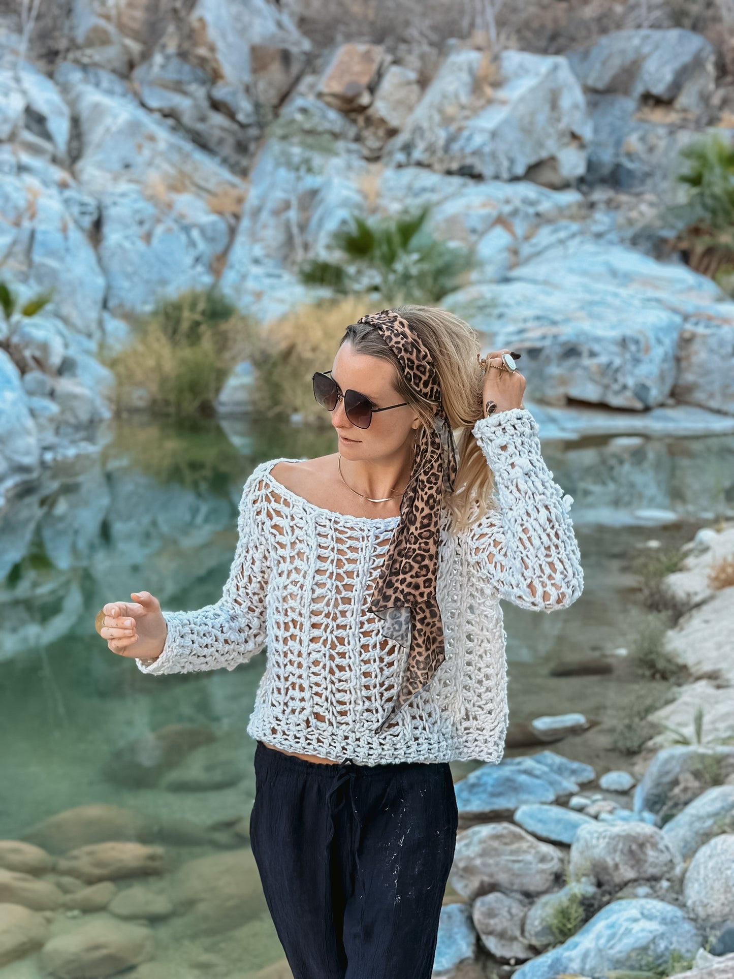 Woman in a white crochet top and black pants standing by a rocky water body.