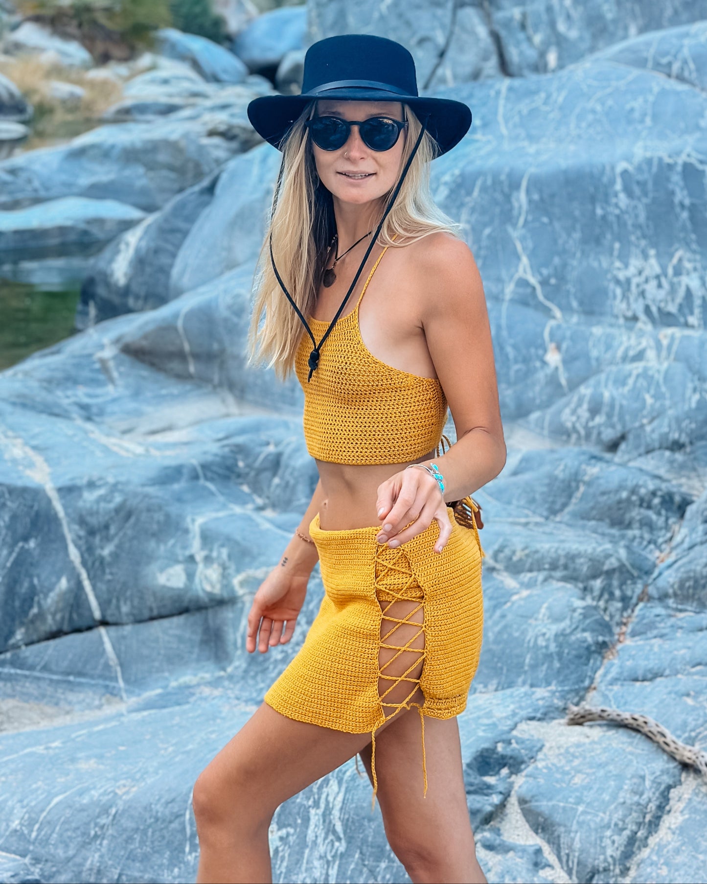 Woman in yellow outfit and black hat standing on rocky terrain with water and trees in the background